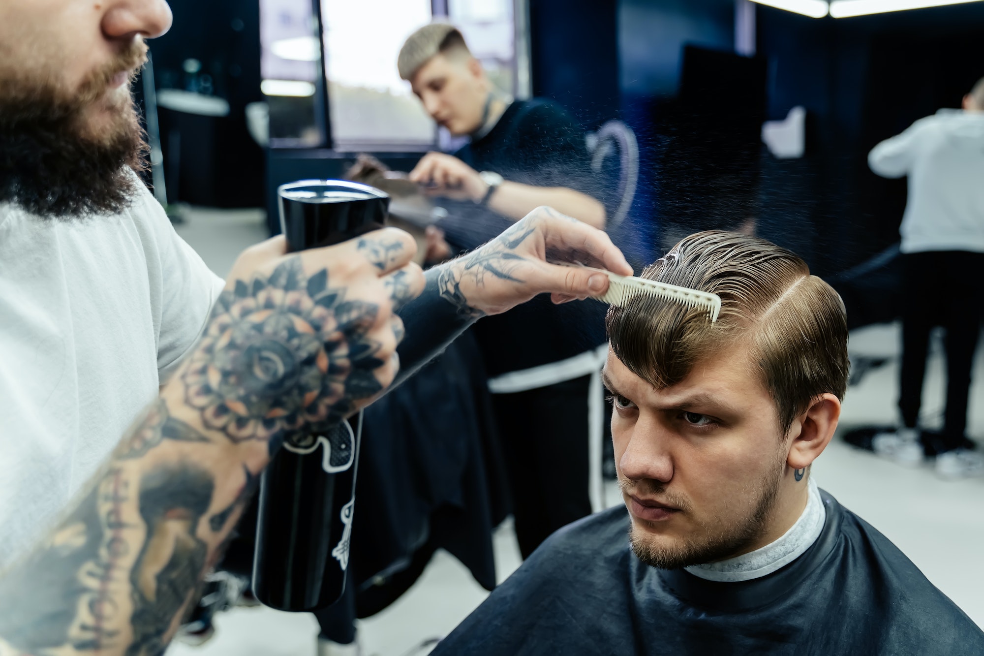 Attractive male is getting a modern haircut in barber shop. Tattooed Barber wets hair by spray and
