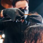 Young bearded man sitting and getting haircut in barber shop by guy in black protective mask