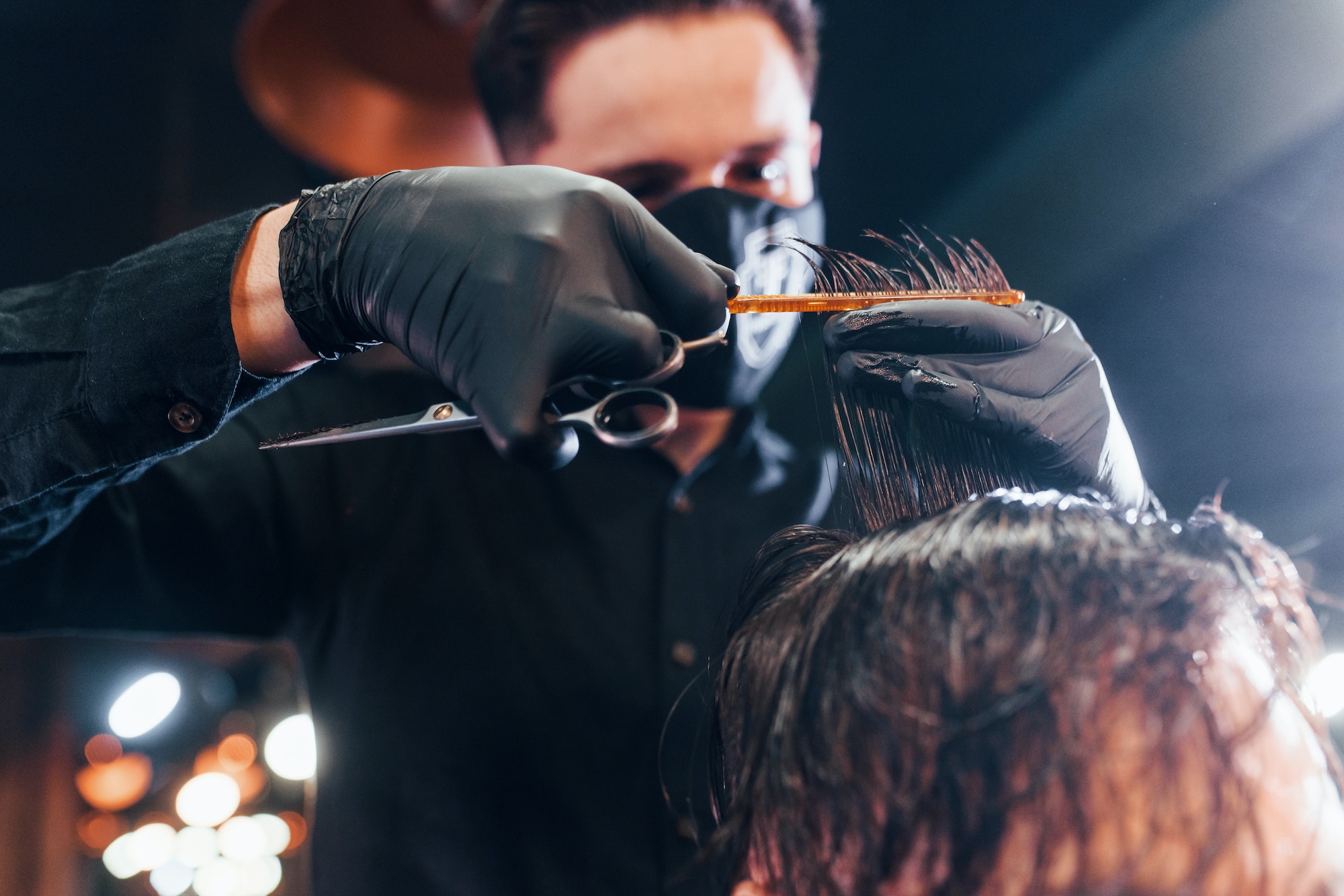 Young bearded man sitting and getting haircut in barber shop by guy in black protective mask
